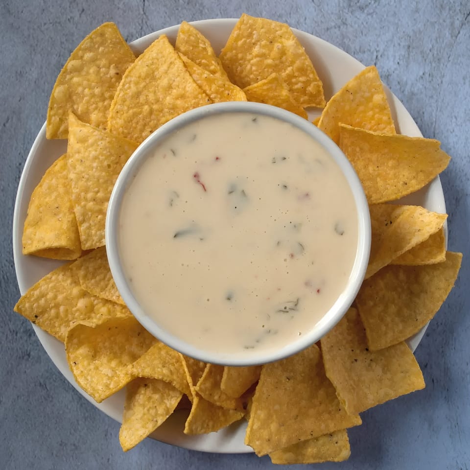 A white bowl of creamy queso dip with specks of green, surrounded by golden tortilla chips.
