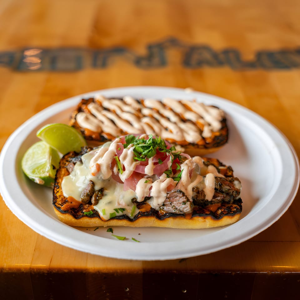 Two Mexican BBQ sandwiches with meat, cheese, sauce, pickled onions, and cilantro, served with lime wedges.
